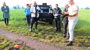 Staatssekretär Gert Zender sowie weitere Menschen stehen auf einem Feld und lassen sich erklären, wie eine Drohne mit Wärmebildtechnologie funktioniert