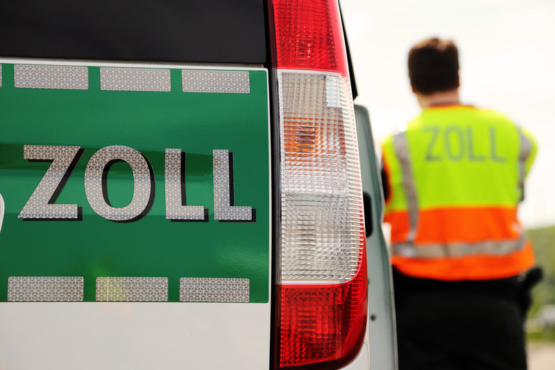 Zoll Fahrzeug mit "Zoll"-Aufschrift, im Hintergrund steht eine Person mit Warnjacke und der Aufschrift "Zoll"